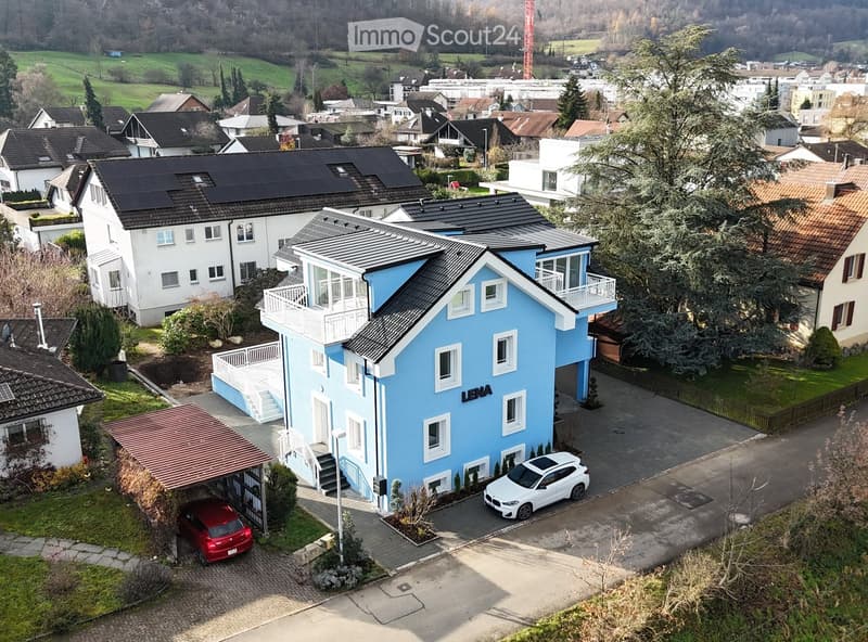Aerial view of a modern two-story house with a blue facade, white trim, and a black roof. The house features a balcony, a driveway with two parked cars, and is surrounded by a well-maintained garden and neighboring houses.