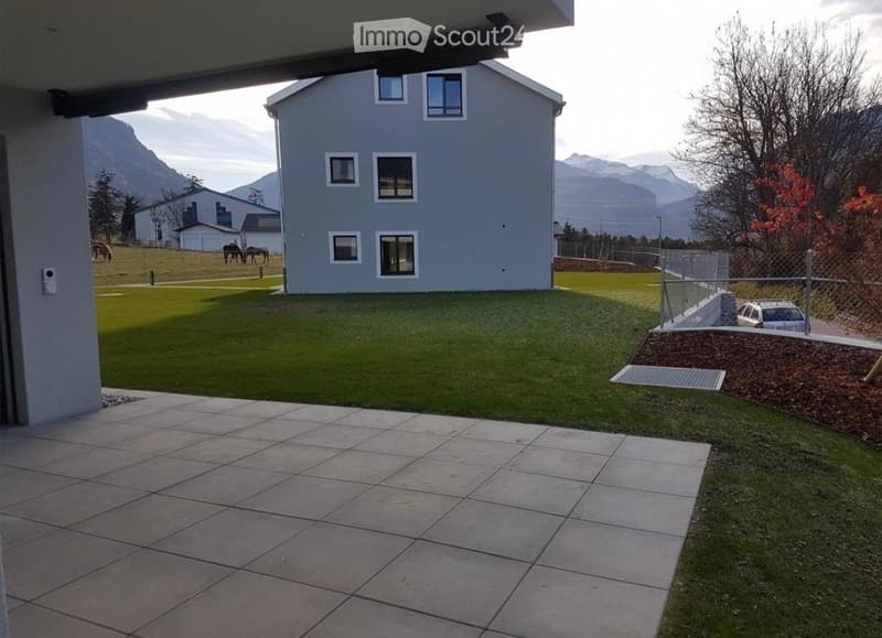 Outdoor patio area with tiled flooring, grassy lawn, and a modern house in the background. The scene includes a fenced area with a parked car and a view of mountains in the distance.