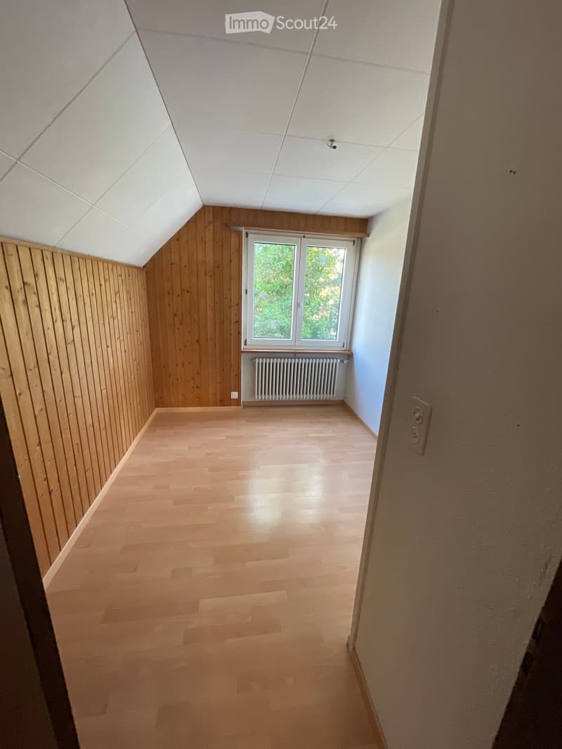 Empty room with wooden walls and floor, large window with view of greenery, radiator below window, and visible electrical outlet on wall.
