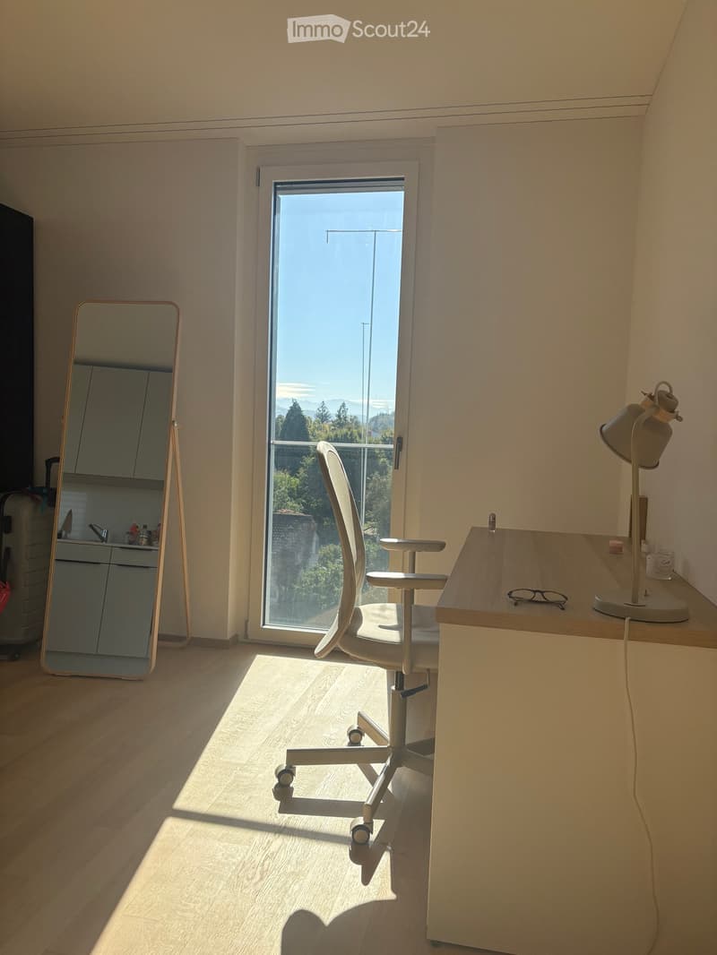 A modern, well-lit room with a wooden floor, a large window offering a scenic view, a white ergonomic chair, a wooden desk with a lamp, and a full-length mirror.
