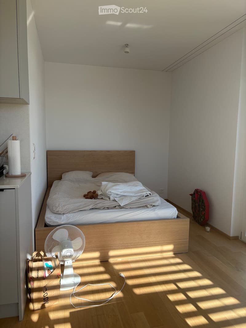 Cozy bedroom with a wooden bed, white bedding, a small fan on the floor, and a paper towel roll on a shelf. Sunlight streams in, casting shadows on the wooden floor.