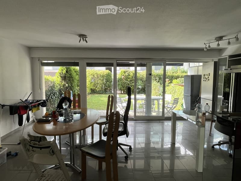 Dining area with a round wooden table, several chairs, and a view of a garden through large sliding glass doors. There is also a refrigerator and a mirror visible.