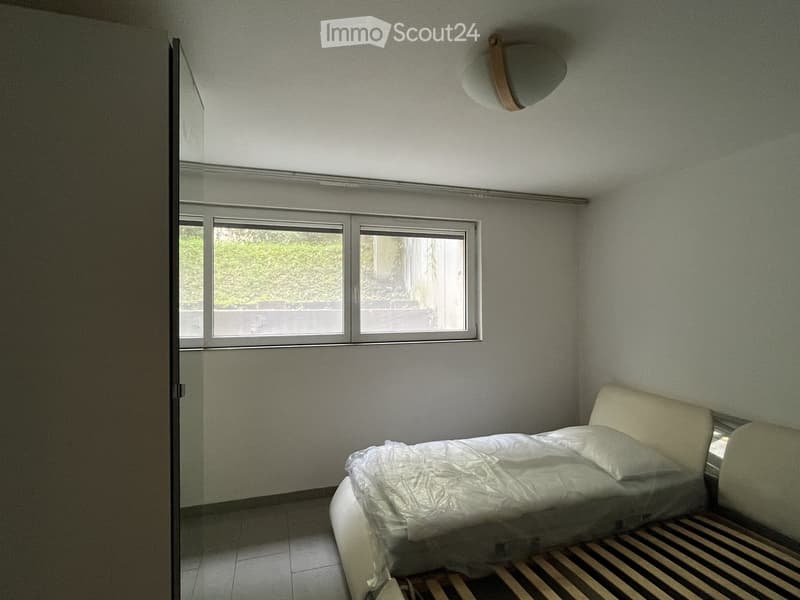 A small, unfurnished bedroom with a single bed, a window, and a radiator. The bed is covered with a plastic sheet, and the room has light-colored walls and a tiled floor.