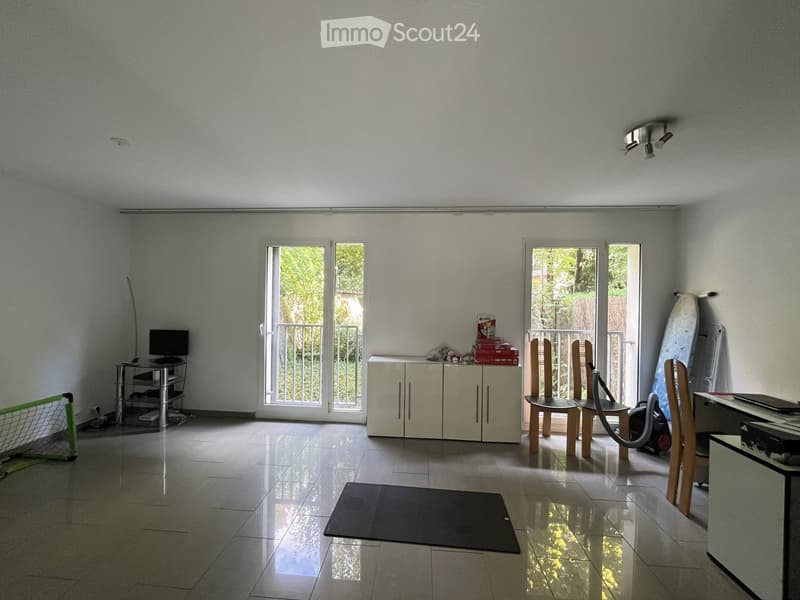 Bright room with tiled floor, large windows, and a balcony. Contains a cabinet, chairs, a screen, a surfboard, and a vacuum cleaner.