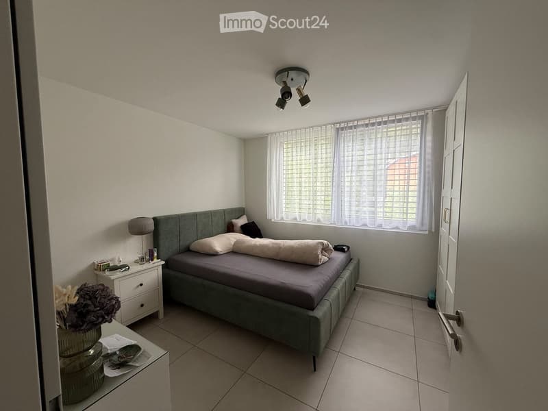 A modern bedroom with a grey upholstered bed, nightstand, lamp, and decorative vase on a small table. The room features tiled flooring and large windows with blinds.