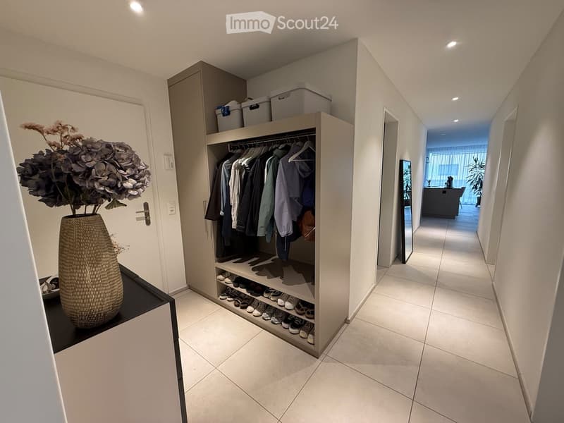 Modern hallway with tiled flooring, built-in closet with clothing and shoes, vase with flowers on a small table, and a view into an adjacent room with a kitchen area.