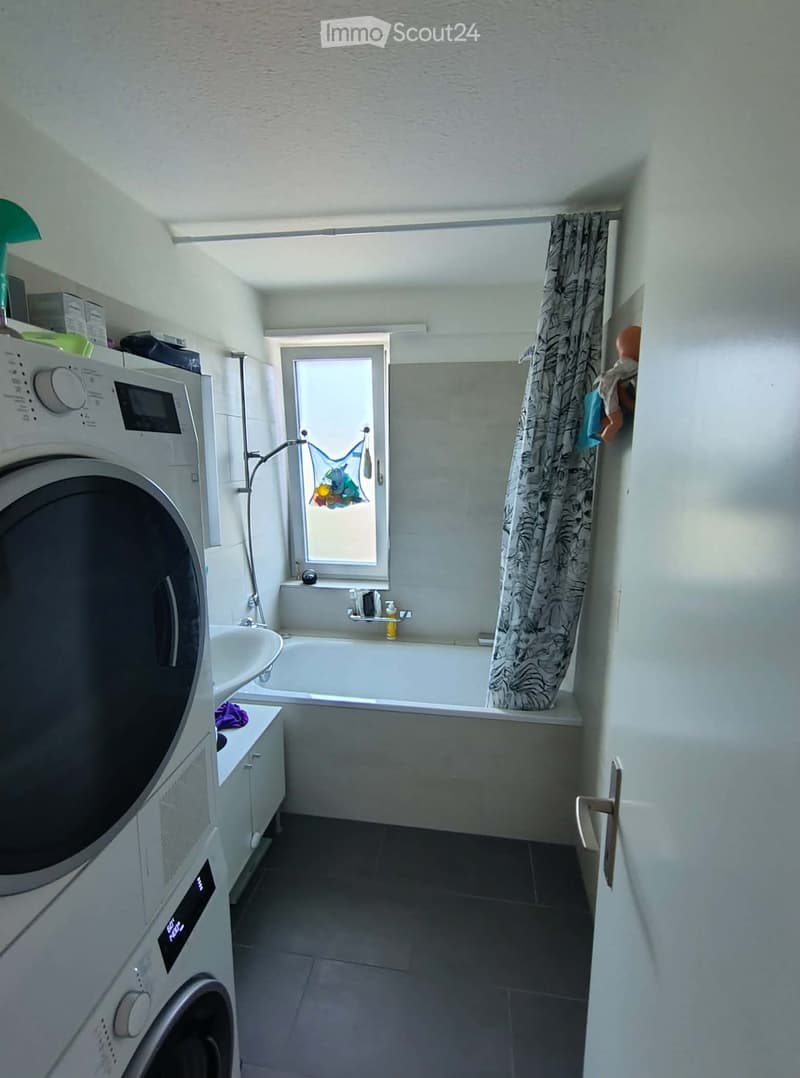 Bathroom with a bathtub, sink, washing machine, and a window with a curtain. The space is well-lit with a skylight and has a modern design.