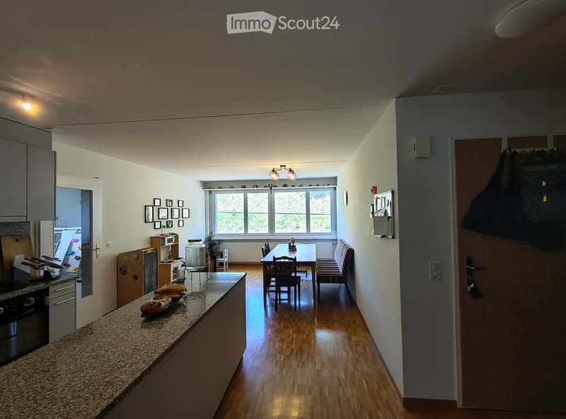 Open-plan kitchen and dining area with wooden flooring, granite countertop, and large window overlooking greenery.