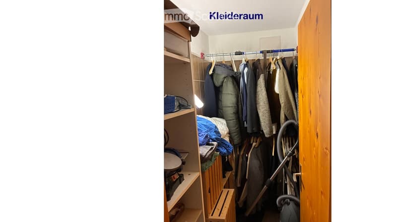 A well-organized closet with wooden shelves, a hanging rod with various garments, and a vacuum cleaner placed on the floor.