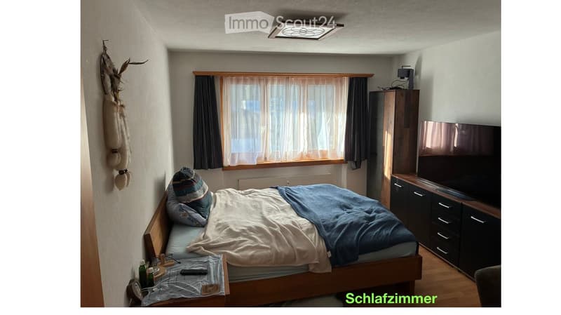 Cozy bedroom with a wooden bed, nightstand, television, and cabinet. The room features a window with curtains and a decorative wall hanging.