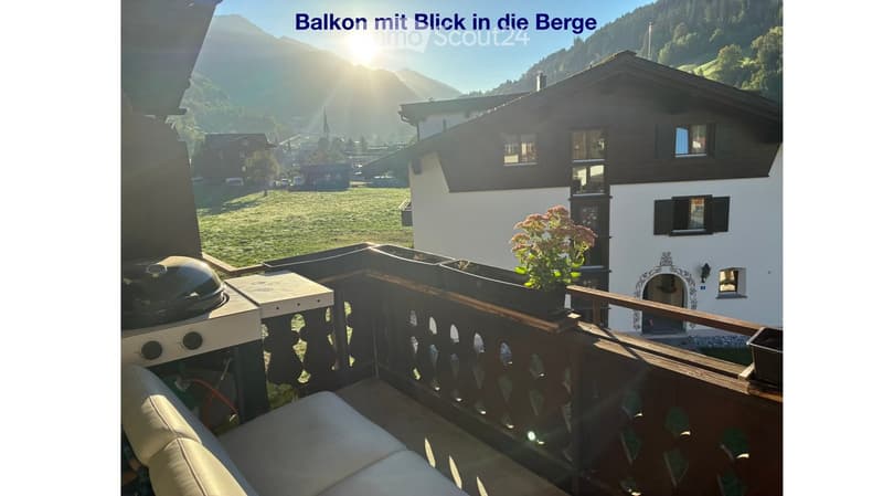 Balcony with wooden railing, sofa, grill, and flower pot, overlooking a scenic mountain view and village.