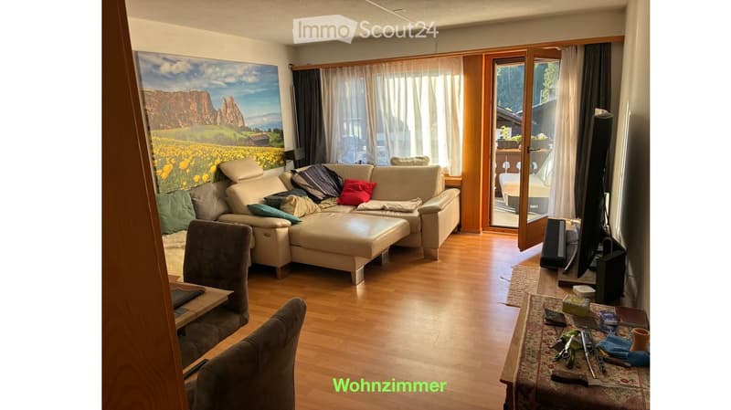 A modern living room with a large sectional sofa, ottoman, and a TV set. The room features wooden flooring, large windows with curtains, and a scenic landscape painting on the wall.