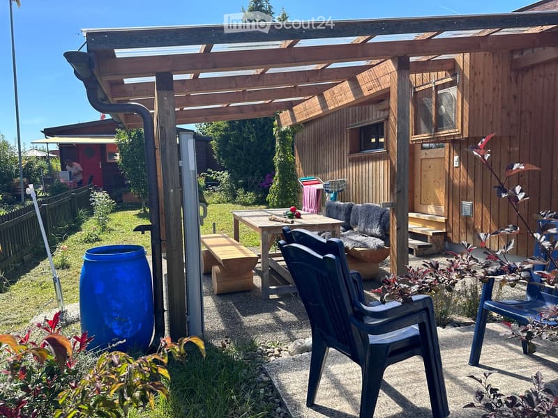 A wooden pergola-covered outdoor area with a table, chairs, a bench, and a large blue barrel. The area is surrounded by plants and a wooden building.