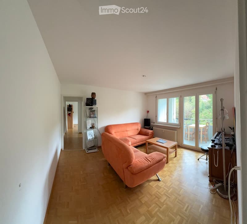 Bright living room with wooden flooring, large windows, and a balcony. Features a pink sofa, coffee table, and a small workspace area with a chair and table.