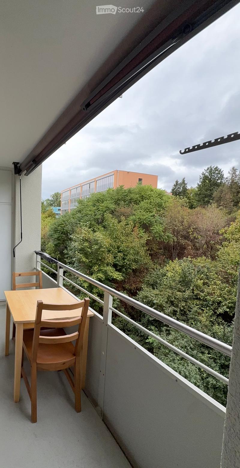 Balcony with a small wooden table and two chairs, overlooking a green area with trees and a building in the background.