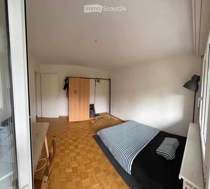 A small, furnished bedroom with a wooden floor, a single bed with striped bedding, a wooden chair, a small table, a wardrobe, and a floor lamp near the window.