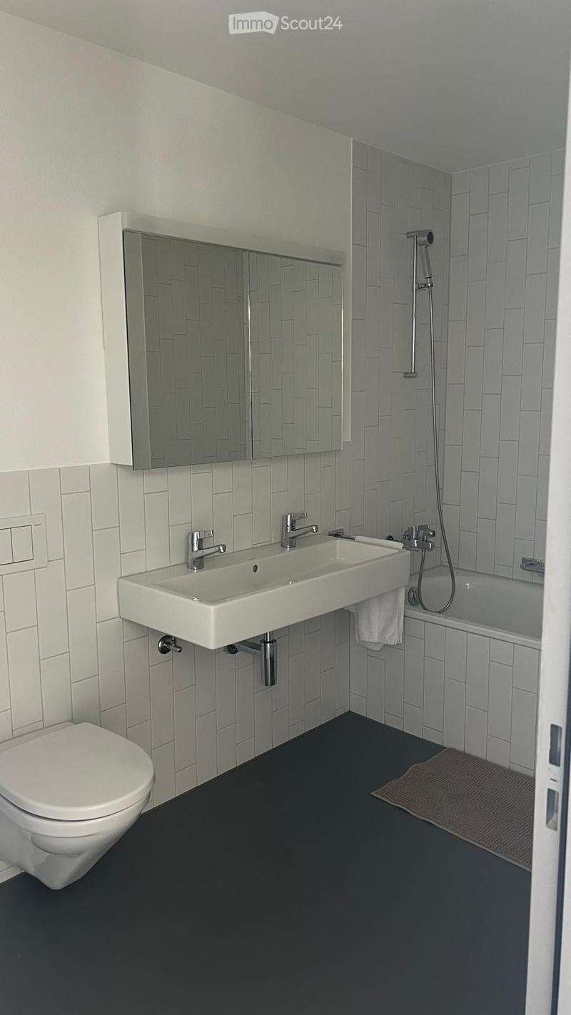 Modern bathroom with a double sink, mirror, toilet, bathtub, and showerhead. The walls are tiled in a light grey pattern, and the floor is dark grey.