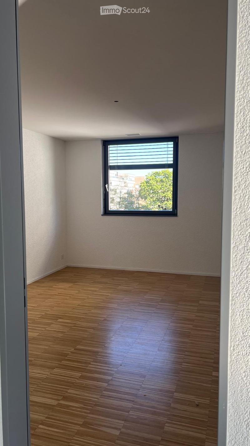 Empty room with wooden flooring and a large window with blinds, showing a view of trees and buildings outside.