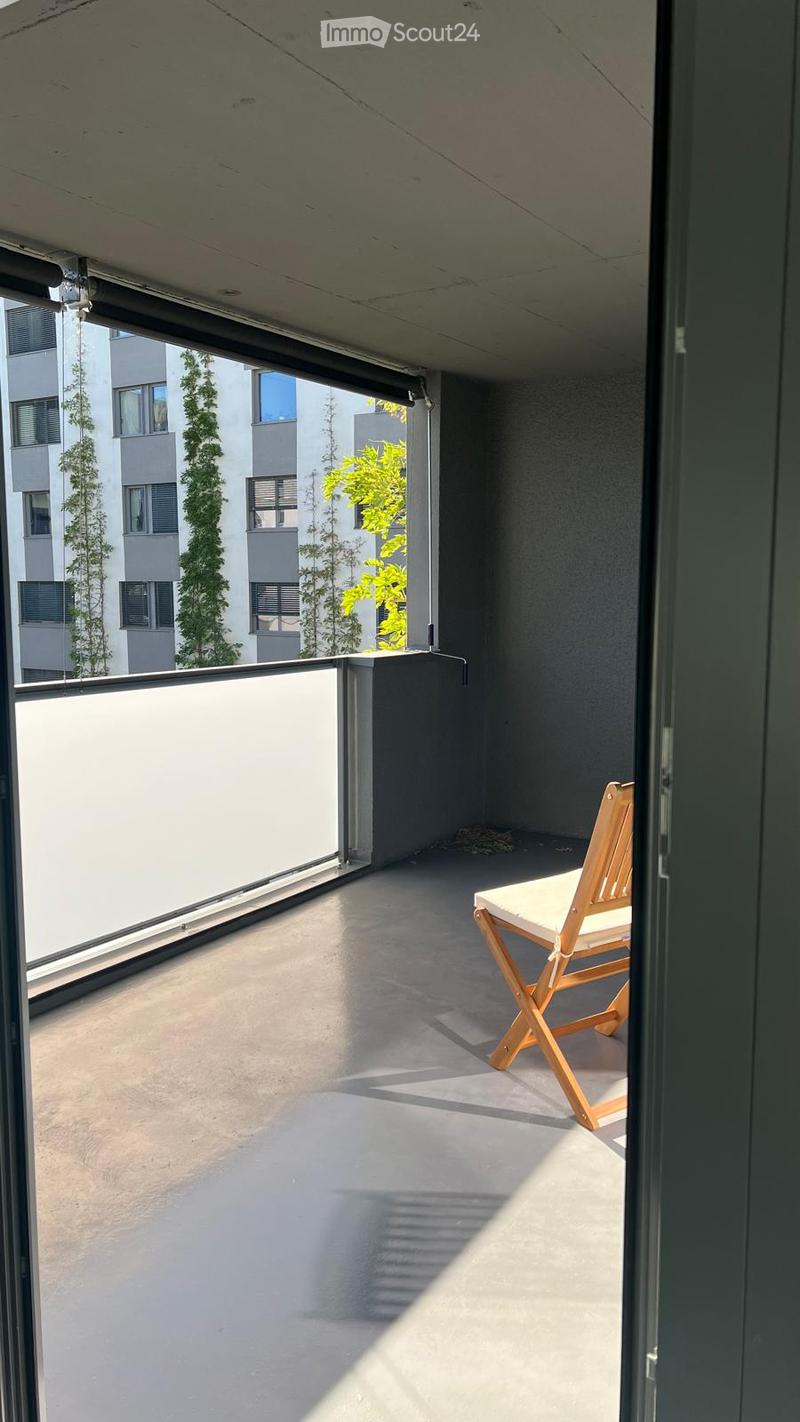 Modern balcony with concrete floor, white table, wooden folding chair, and a view of neighboring buildings and greenery.