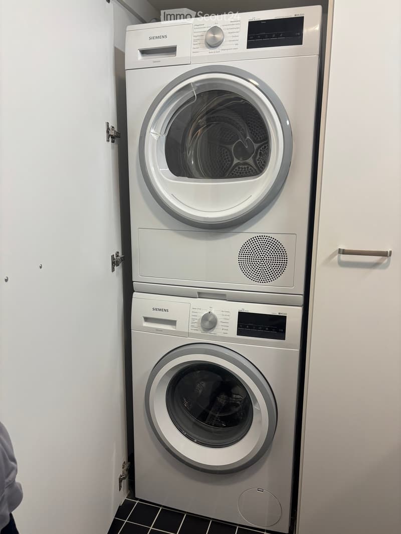 Modern bathroom with a stacked Siemens washing machine and dryer unit, white tiled walls, and black tiled floor.