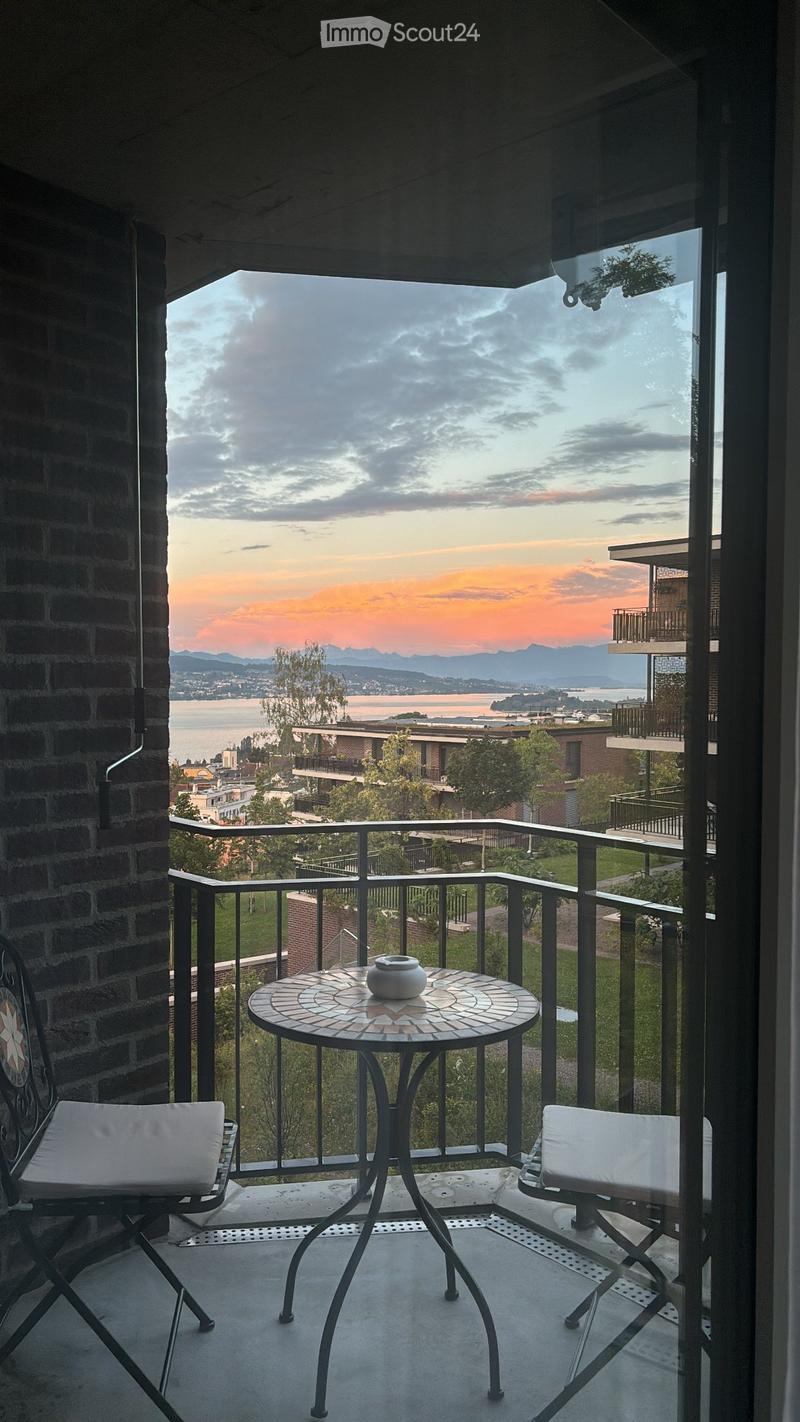 Balcony with a round mosaic table, two white cushioned chairs, and a decorative vase, overlooking a scenic view of a lake and distant mountains.
