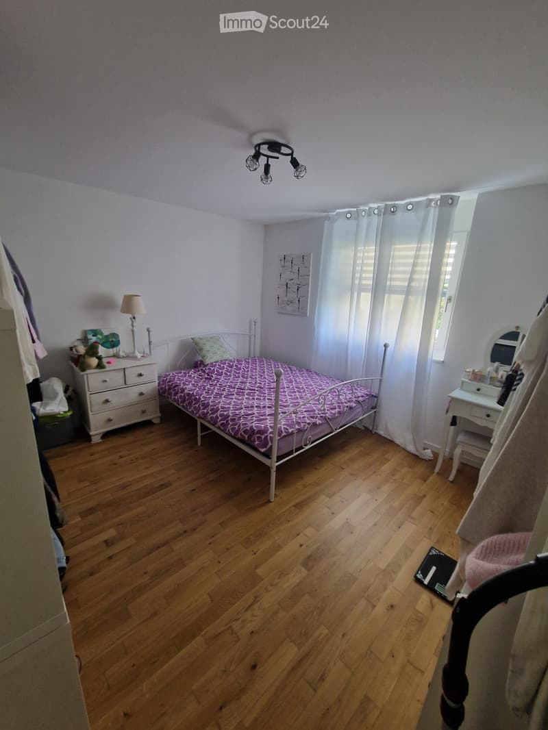 Cozy bedroom with wooden flooring, a single bed with a purple patterned bedspread, a nightstand with a lamp, a mirror on a small table, and a small stool near the window.