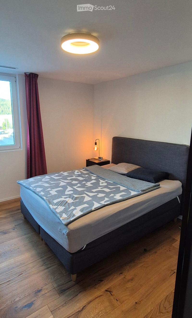Modern bedroom with a large bed, nightstand, and a round ceiling light. The room features wooden flooring and a window with a curtain.