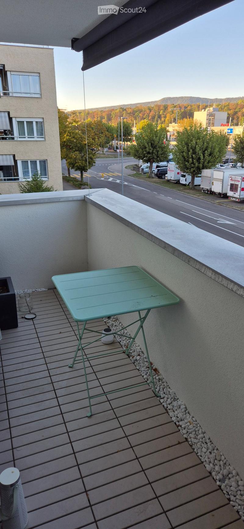 Balcony with wooden decking, green metal table, small potted plant, and a view of the street and surrounding area.
