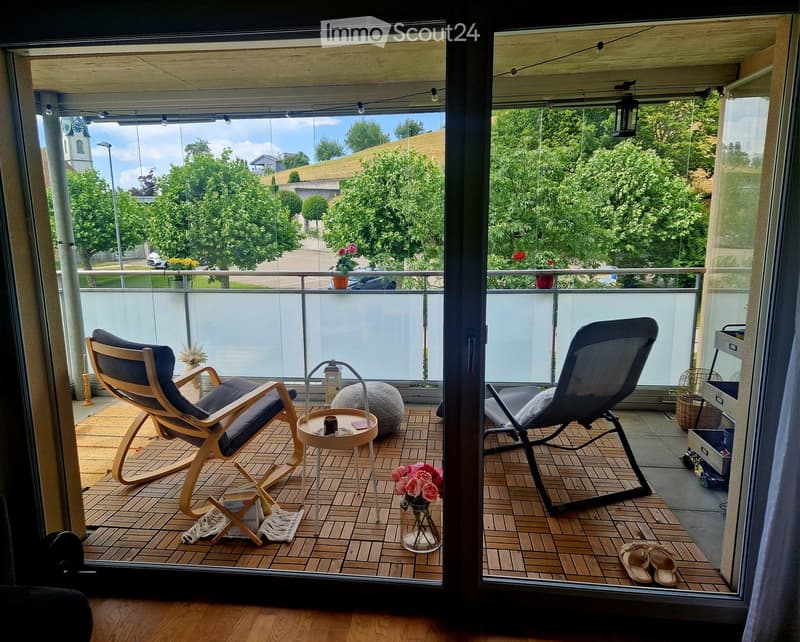 Balcony with wooden decking, two chairs, a small table, a round stool, and a flower vase with pink roses.