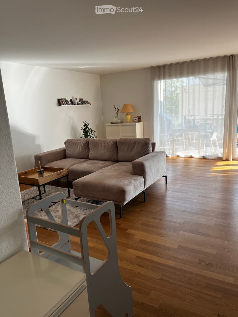 Modern living room with a grey sectional sofa, wooden coffee table, small chair, table lamp, and a shelf with decorative items. The room features wooden flooring and large windows with sheer curtains.