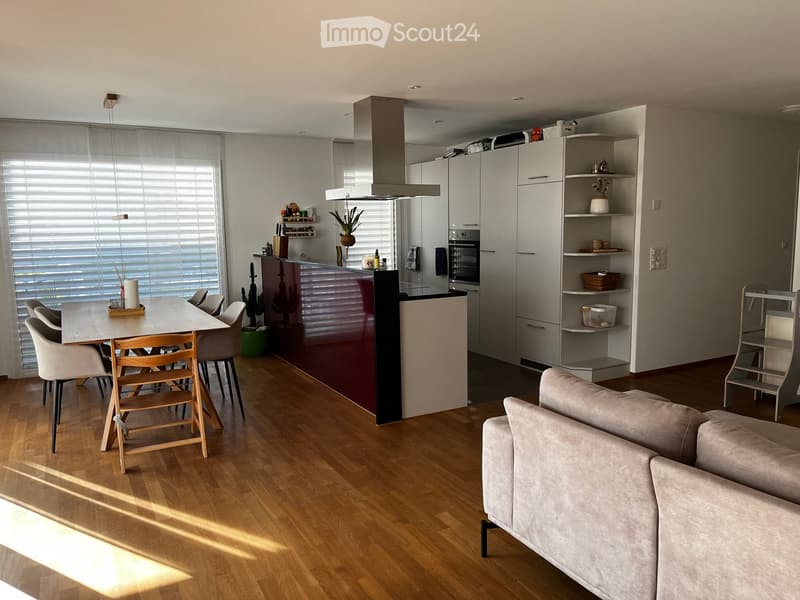 Open-plan living and dining area with a modern kitchen, featuring a light grey sofa, a wooden dining table with chairs, and white kitchen cabinets with integrated appliances.