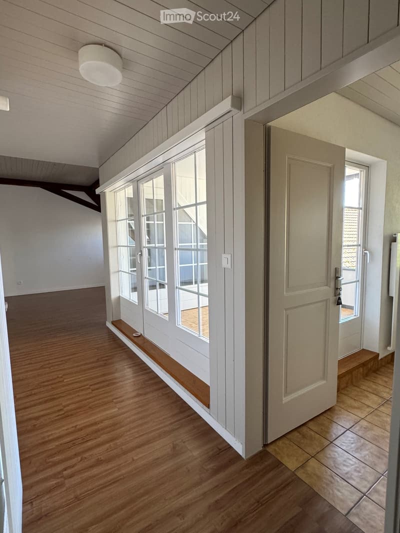 Bright hallway with wooden flooring, white walls, and large windows. Features a built-in wooden bench and a closed door leading to another room.