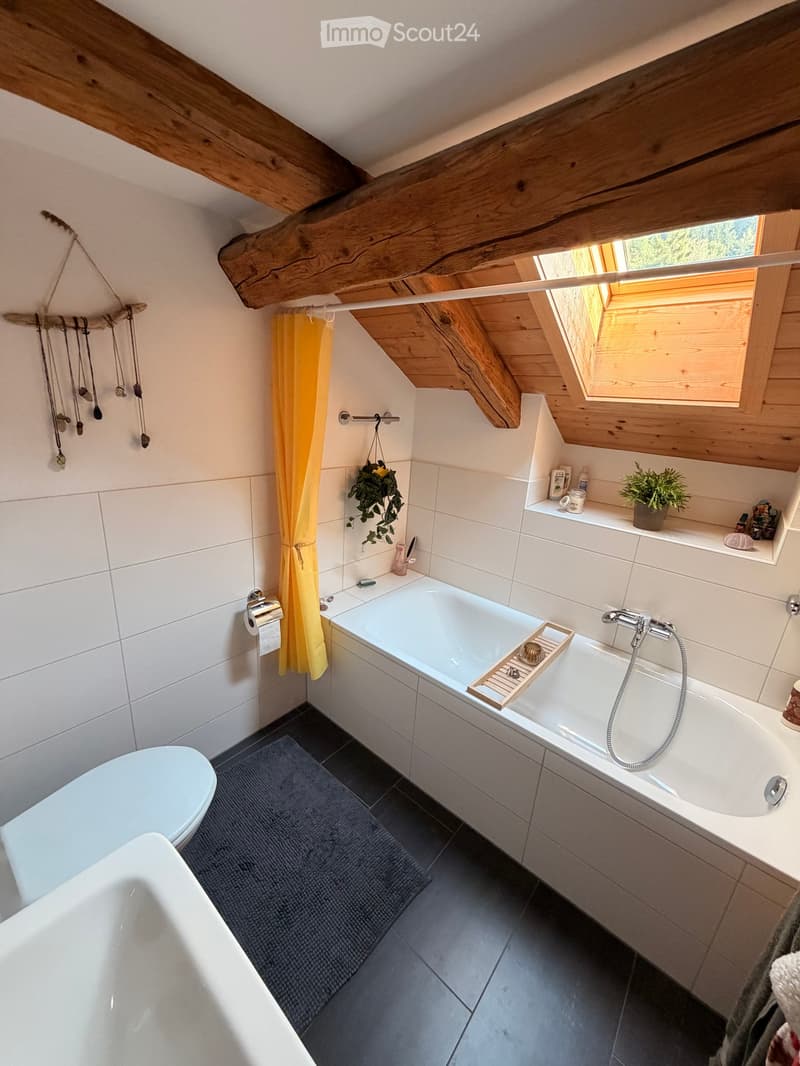 Cozy bathroom with exposed wooden beams, a bathtub with a shower curtain, a toilet, a towel, a small plant, and a shelf with toiletries.