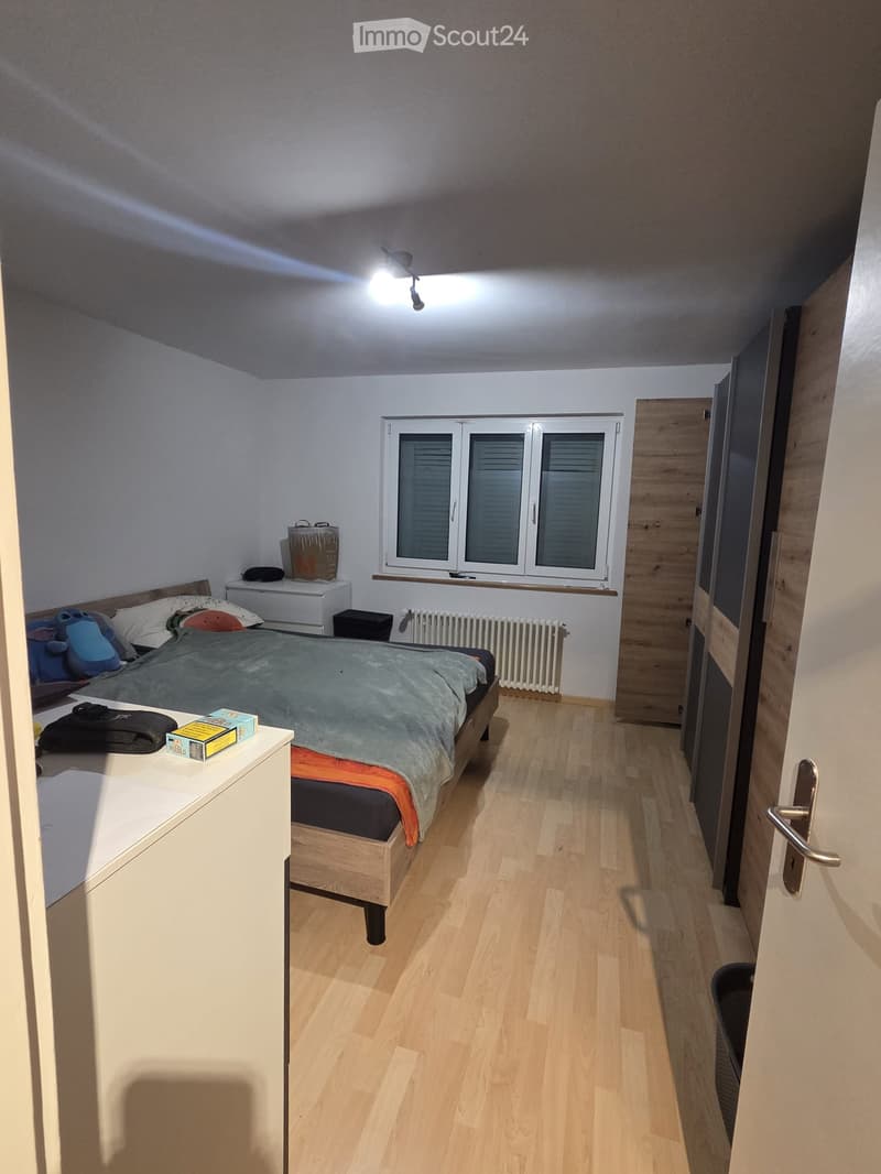Cozy bedroom with wooden flooring, a double bed with pillows and blankets, a nightstand with a lamp, a window with blinds, and a radiator below it.