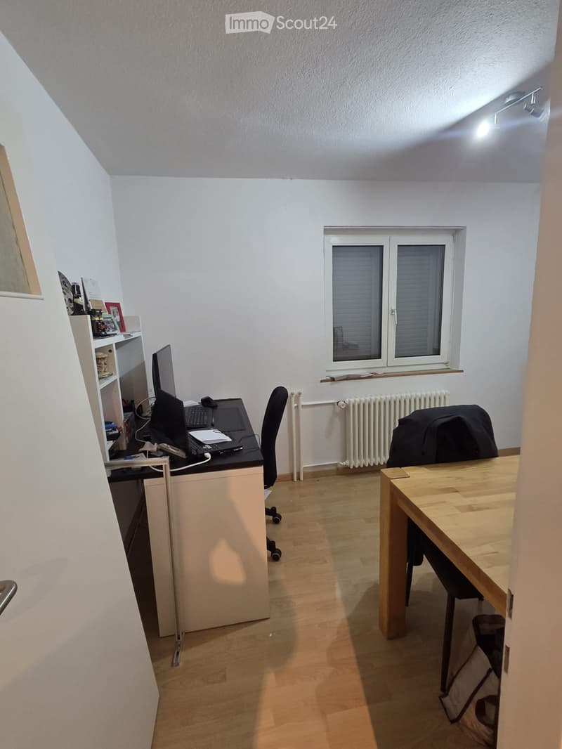 A well-lit room with wooden flooring, a desk with a laptop and chair, a wooden table, a shelf with various items, and a window with blinds.