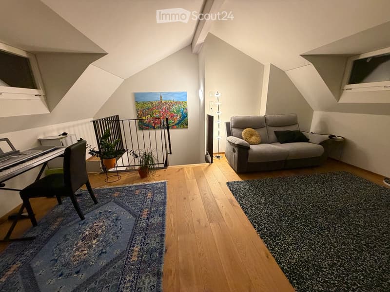 Cozy attic living room with wooden flooring, a grey sofa, a piano, a chair, a small table, potted plants, a colorful painting, and two rugs.