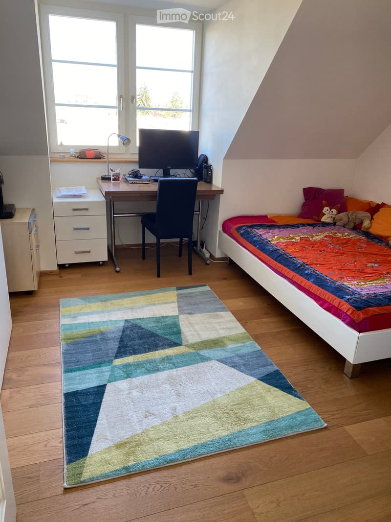 Bright bedroom with a bed, desk, chair, and colorful rug. The room features wooden flooring and large windows letting in natural light.