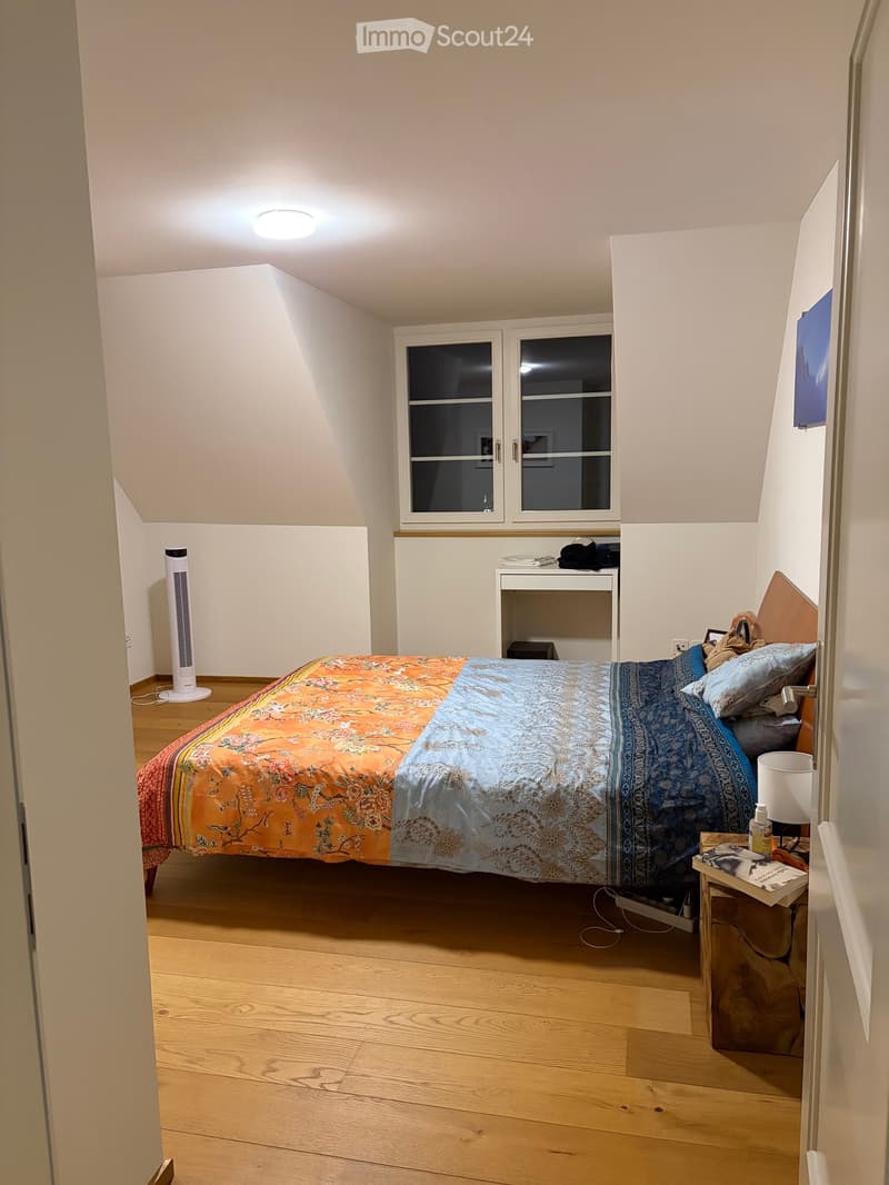 Cozy bedroom with wooden flooring, a large bed with colorful bedding, a nightstand with a lamp, a fan, and a small shelf with items.