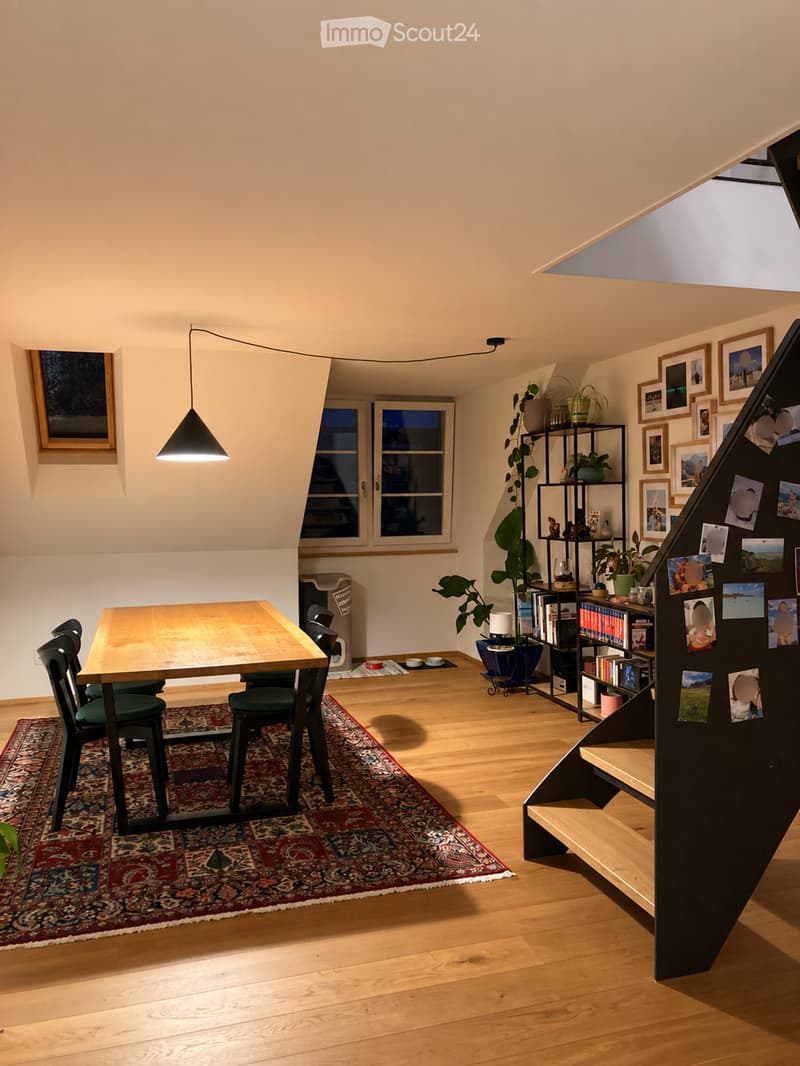 Bright, open room with wooden flooring, a dining table with chairs, a large plant, a bookshelf, and a staircase leading upwards.