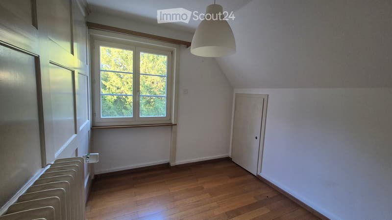 Empty room with wooden flooring, white walls, a window with a view of greenery, a radiator, and a hanging light fixture.