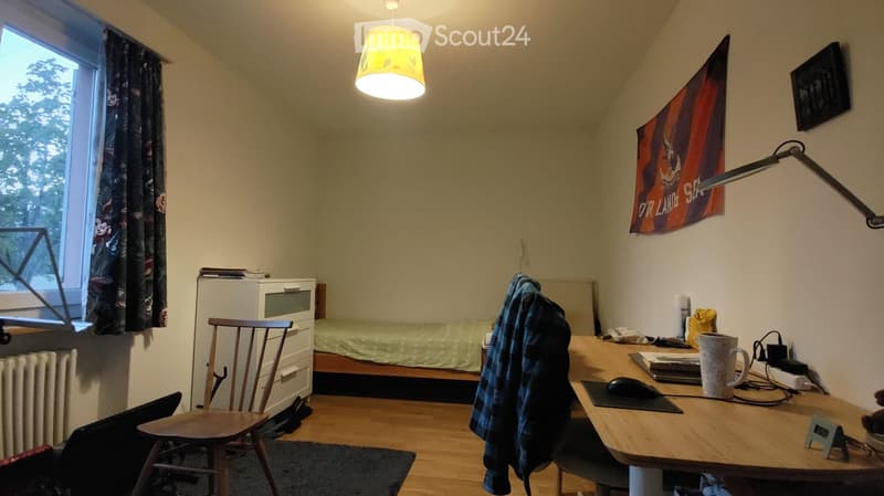 A small, cozy bedroom with a bed, desk, chair, and various personal items. The room features wooden flooring, a window with curtains, and a slanted ceiling typical of an attic space.