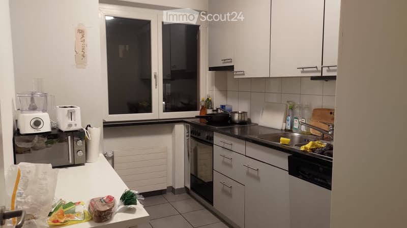 Modern kitchen with white cabinets, stainless steel appliances, and a window. Visible items include a microwave, toaster, food processor, sink, and various kitchen utensils.