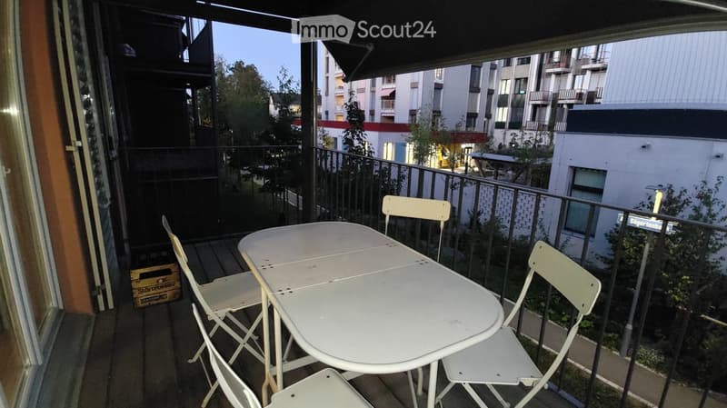 Balcony with a white table and four matching chairs, overlooking a cityscape with buildings and trees.