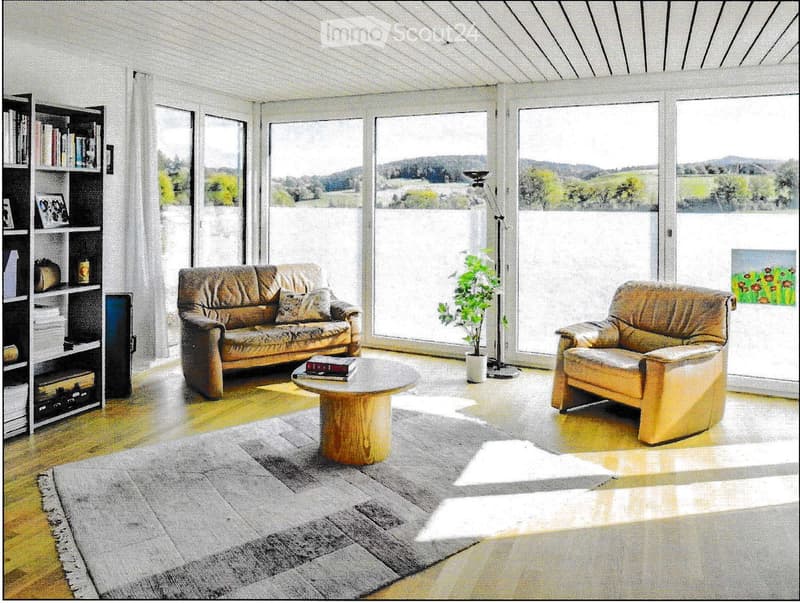 Bright living room with large windows offering a scenic view, featuring a sofa, armchair, coffee table, bookshelf, floor lamp, potted plant, and a painting on the wall.