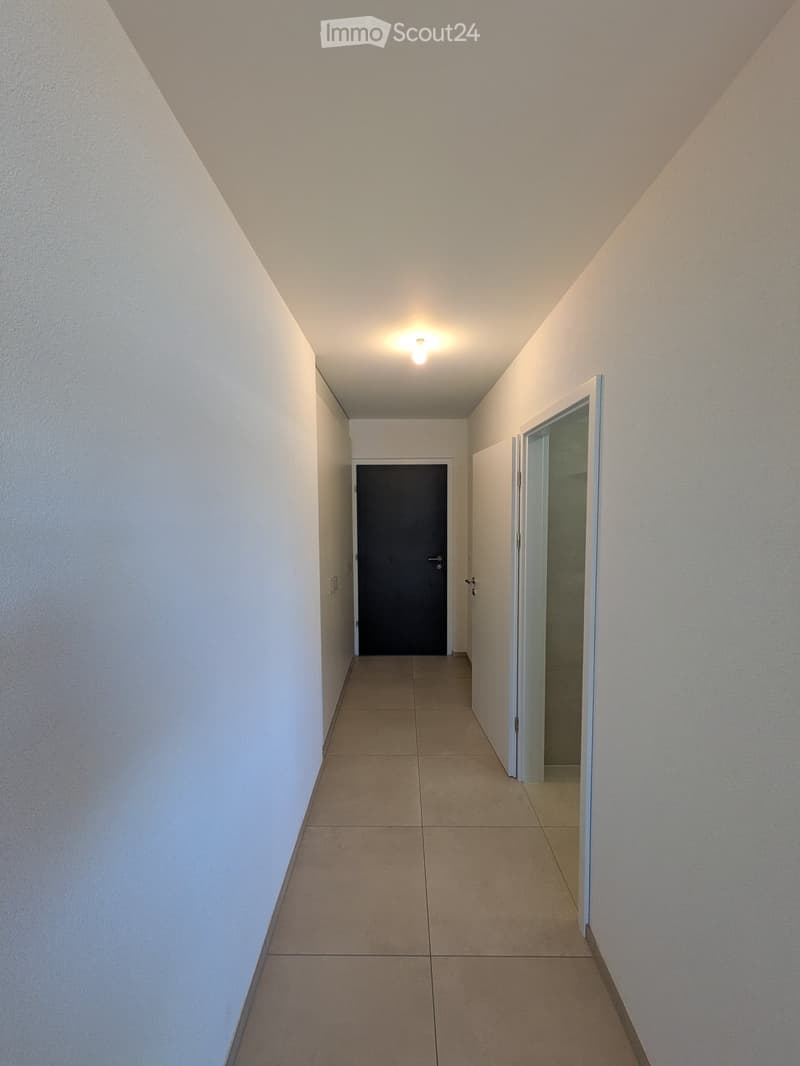 Empty hallway with beige tiled floor, white walls, and a single ceiling light. Features a closed door at the end and an open door on the right side.