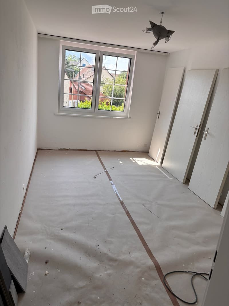 Empty room with bare walls and concrete floor, featuring a large window and several doors.