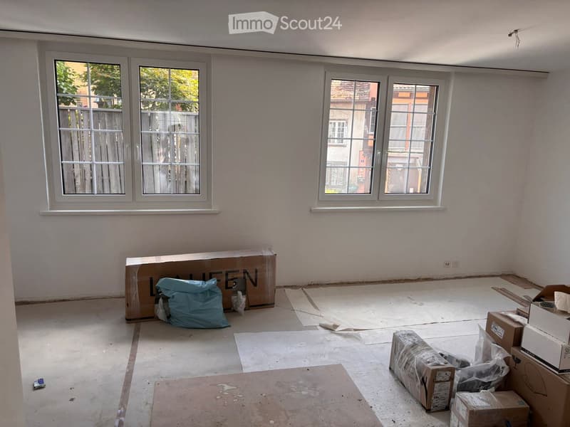 Empty room with white walls, large windows, and scattered boxes and bags on the floor.