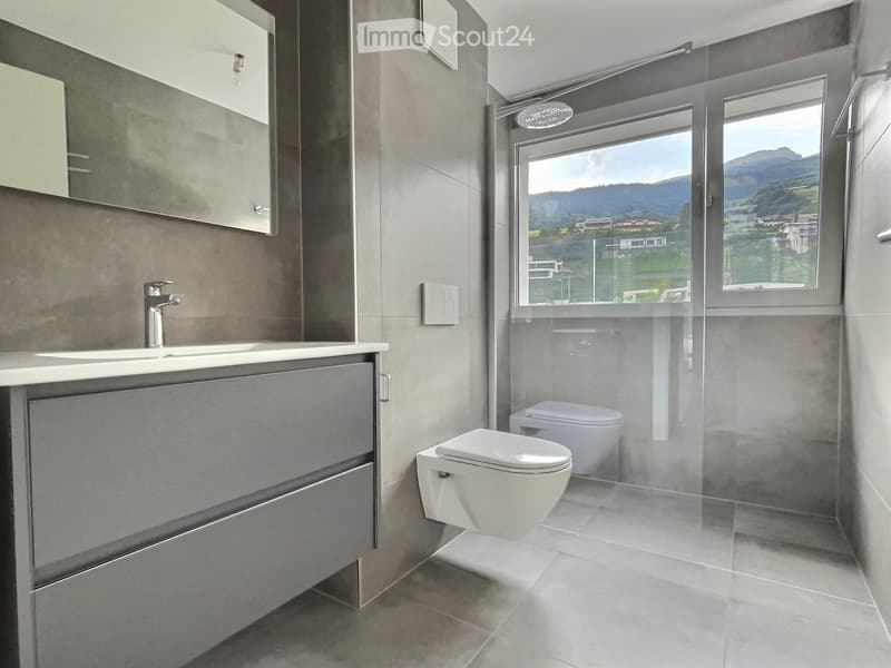 Modern bathroom with a sink, faucet, mirror, cabinet, and toilet, featuring large windows with a scenic mountain view.