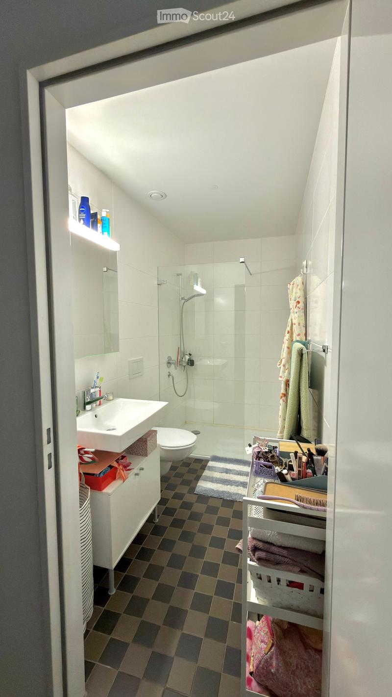 Modern bathroom with white tiles, a glass-enclosed shower, a sink, a toilet, and various personal care items on shelves and a basket.