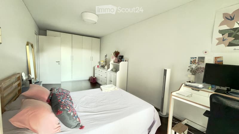 A modern bedroom with a neatly made bed, a desk with a computer, a wardrobe, and decorative items on shelves.